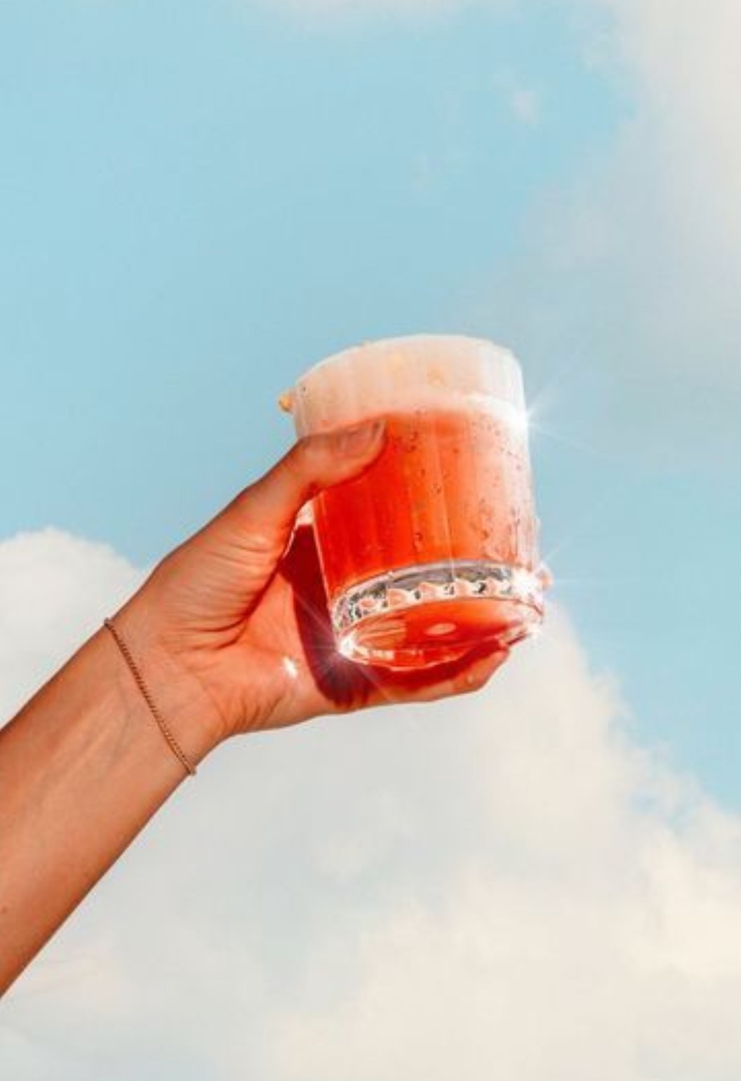 Una mano solleva un bicchiere con una soda rossa contro un cielo azzurro con nuvole.